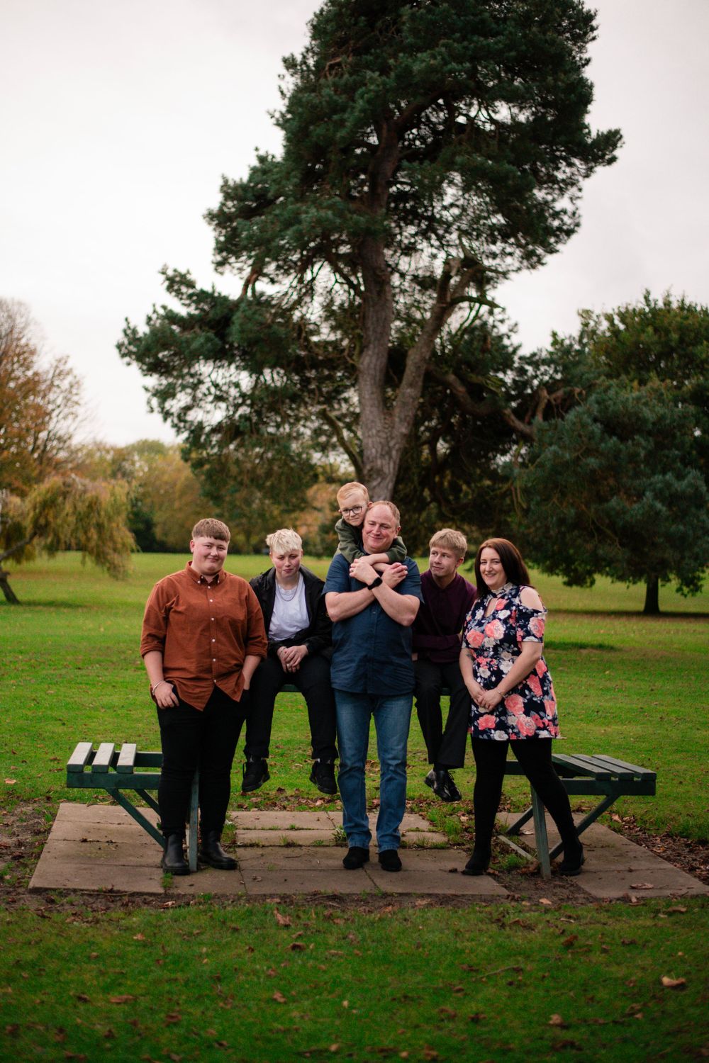 A group poses together on a wooden platform in front of a large pine tree in a park setting.