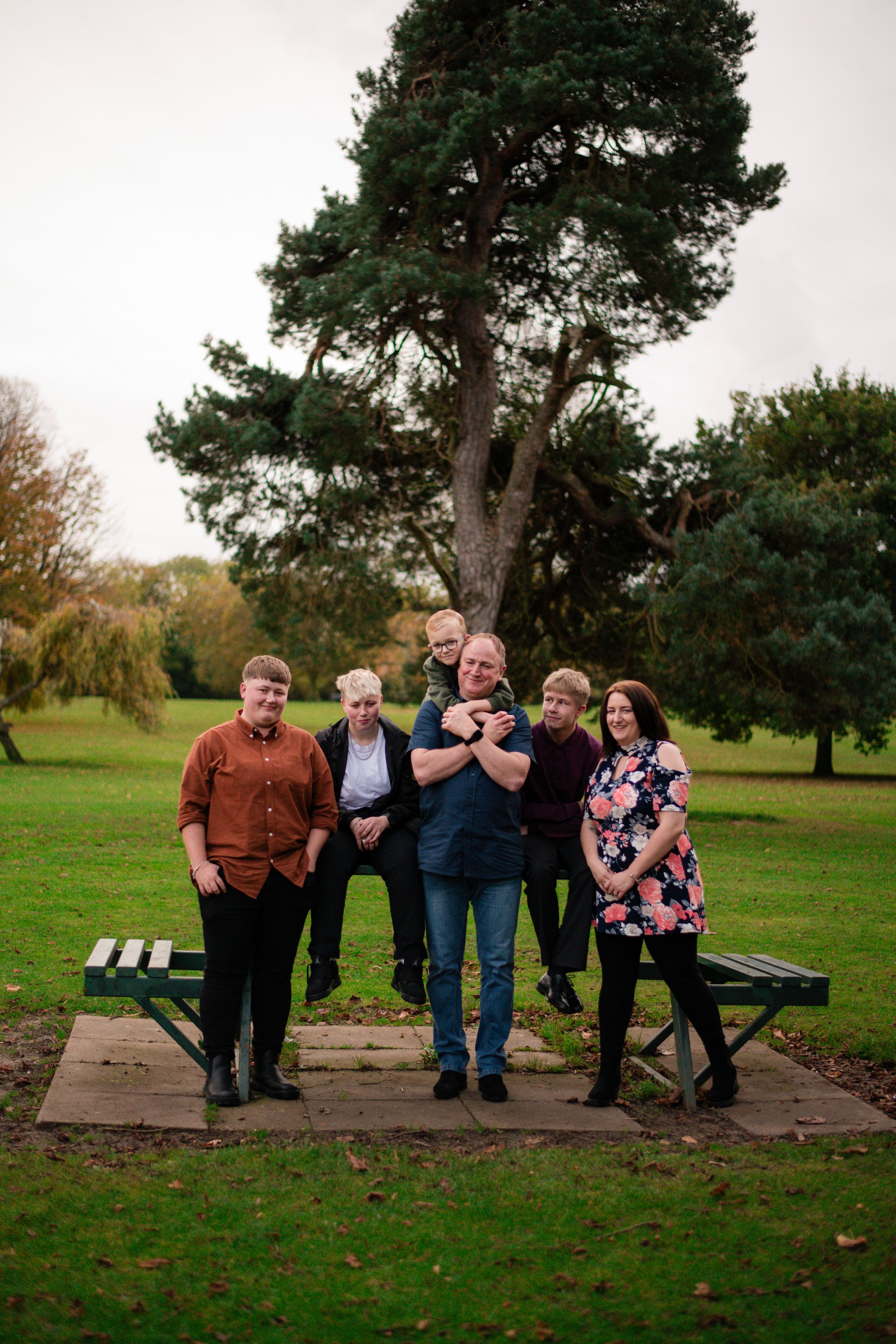 A group poses together on a wooden platform in front of a large pine tree in a park setting.