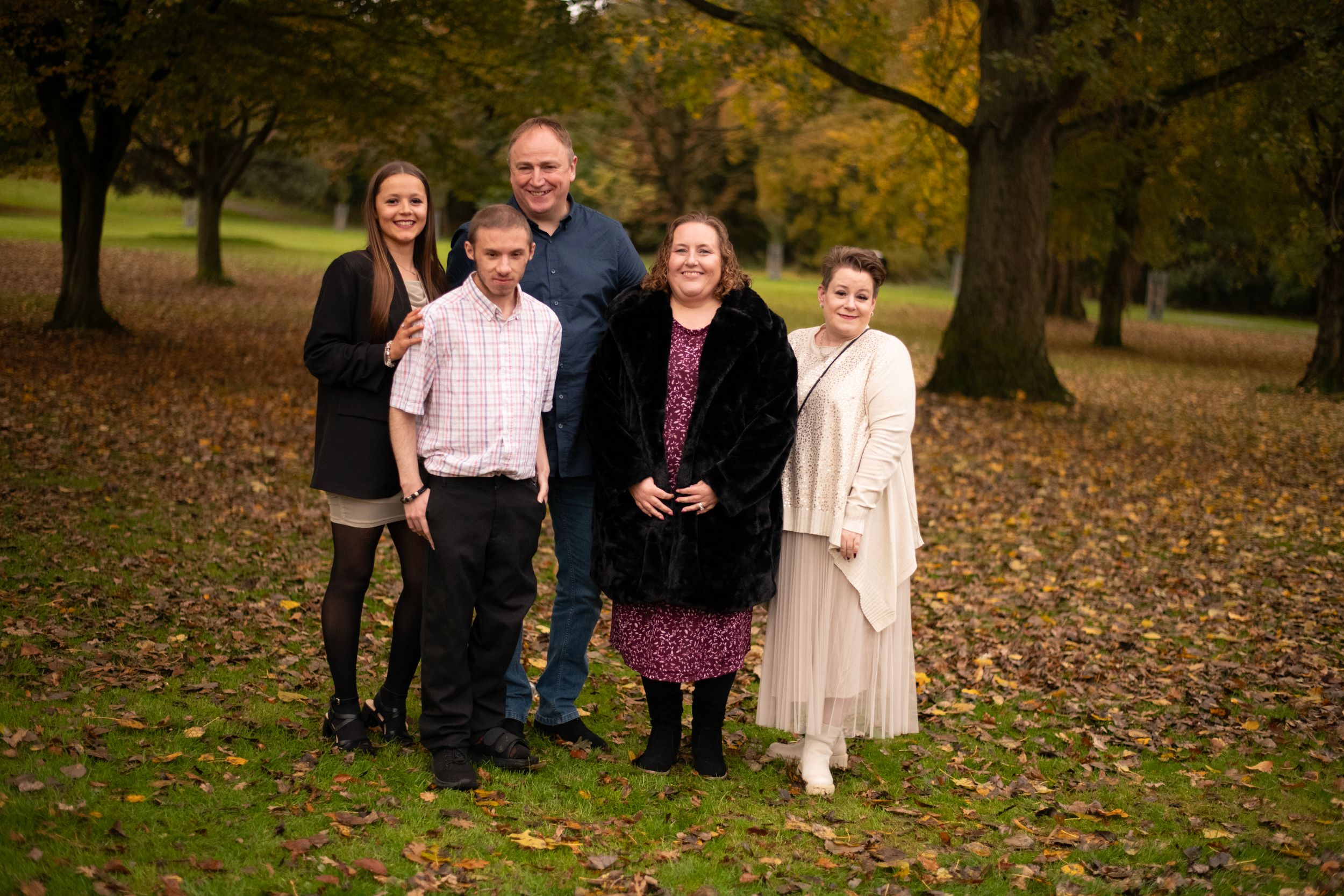 Small family group poses for photo in park with autumn leaves on ground.