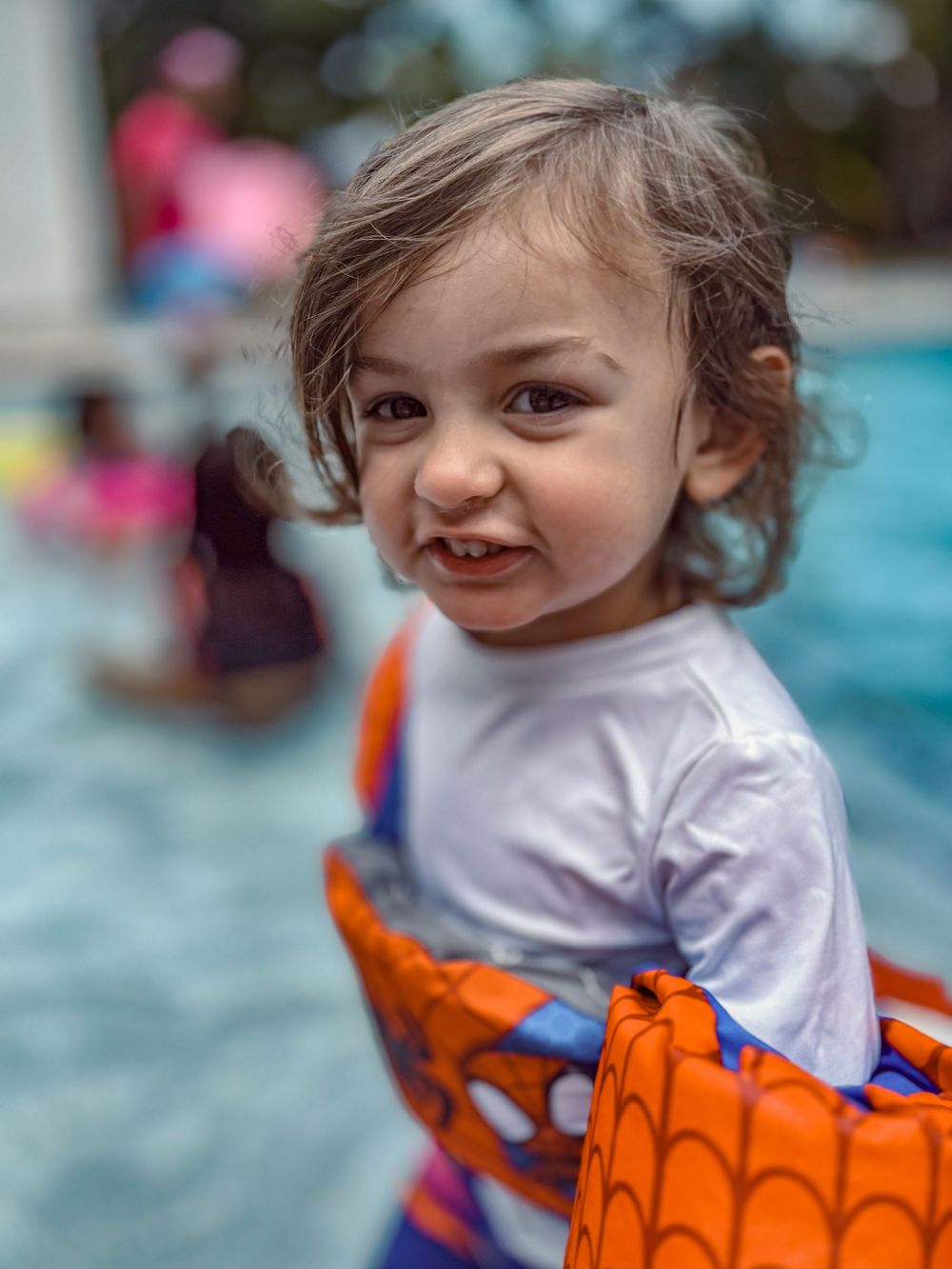 A young child wearing a white shirt and orange floaties by a swimming pool.