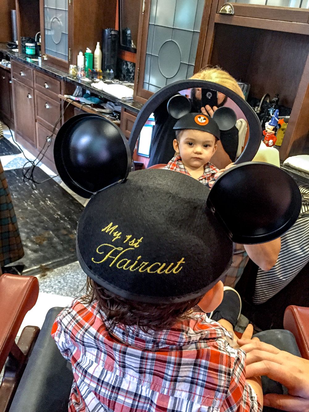 Child getting first haircut wearing Mickey Mouse ears at barbershop.