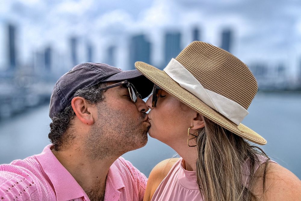 A couple sharing a kiss against a cityscape backdrop near the water.