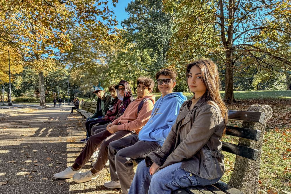 Group of people sitting on park bench surrounded by autumn trees.