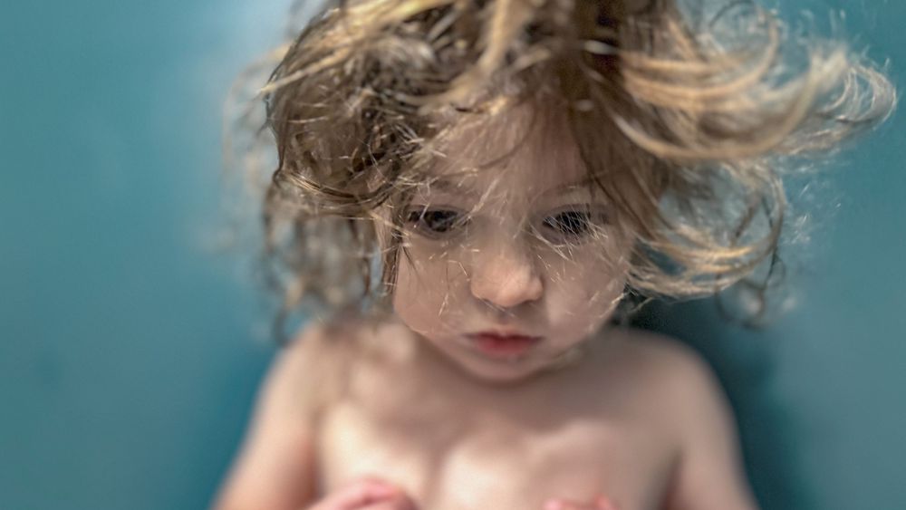 Close-up artistic portrait of a child with windswept hair against turquoise background.