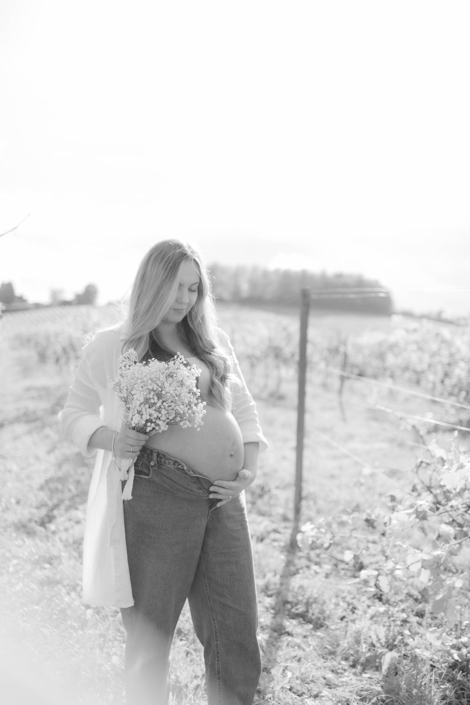 Black and white maternity photo with figure holding flowers in rural setting.