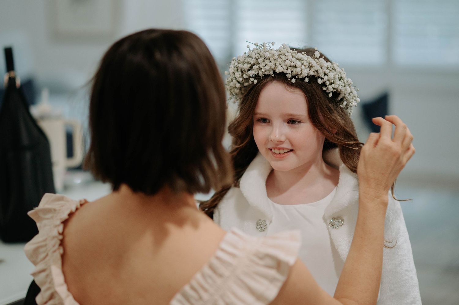 A person wearing a delicate floral crown helps adjust clothing details in a soft, well-lit indoor setting.