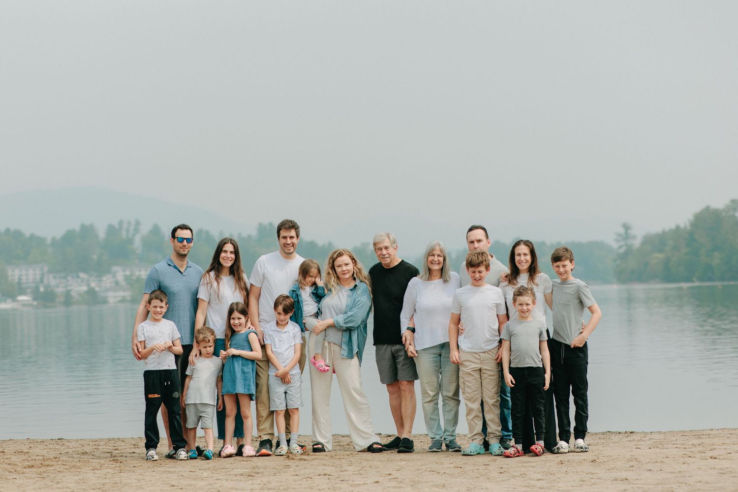 A large family group stands together on a scenic beach with a tranquil lake and forested shore in the background.
