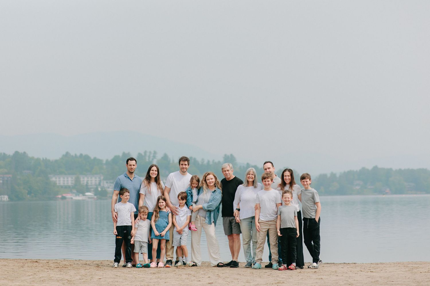A large group stands together on a sandy beach shoreline with a calm lake and trees in the background.