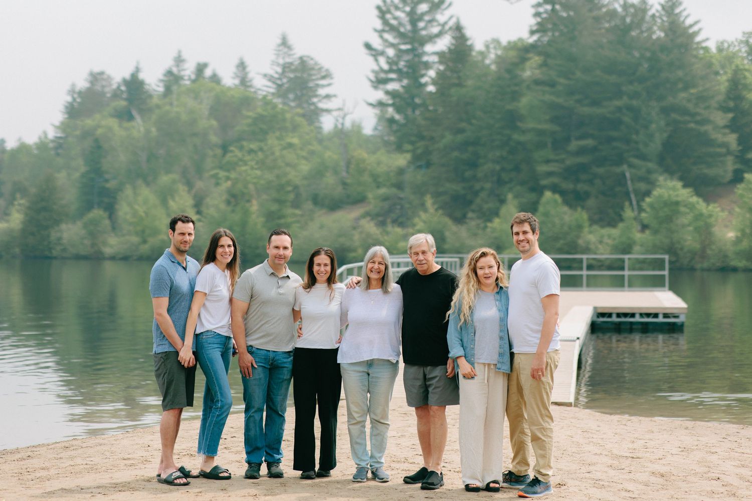A group stands together on a sandy beach by a serene lake surrounded by evergreen trees on a summer day.
