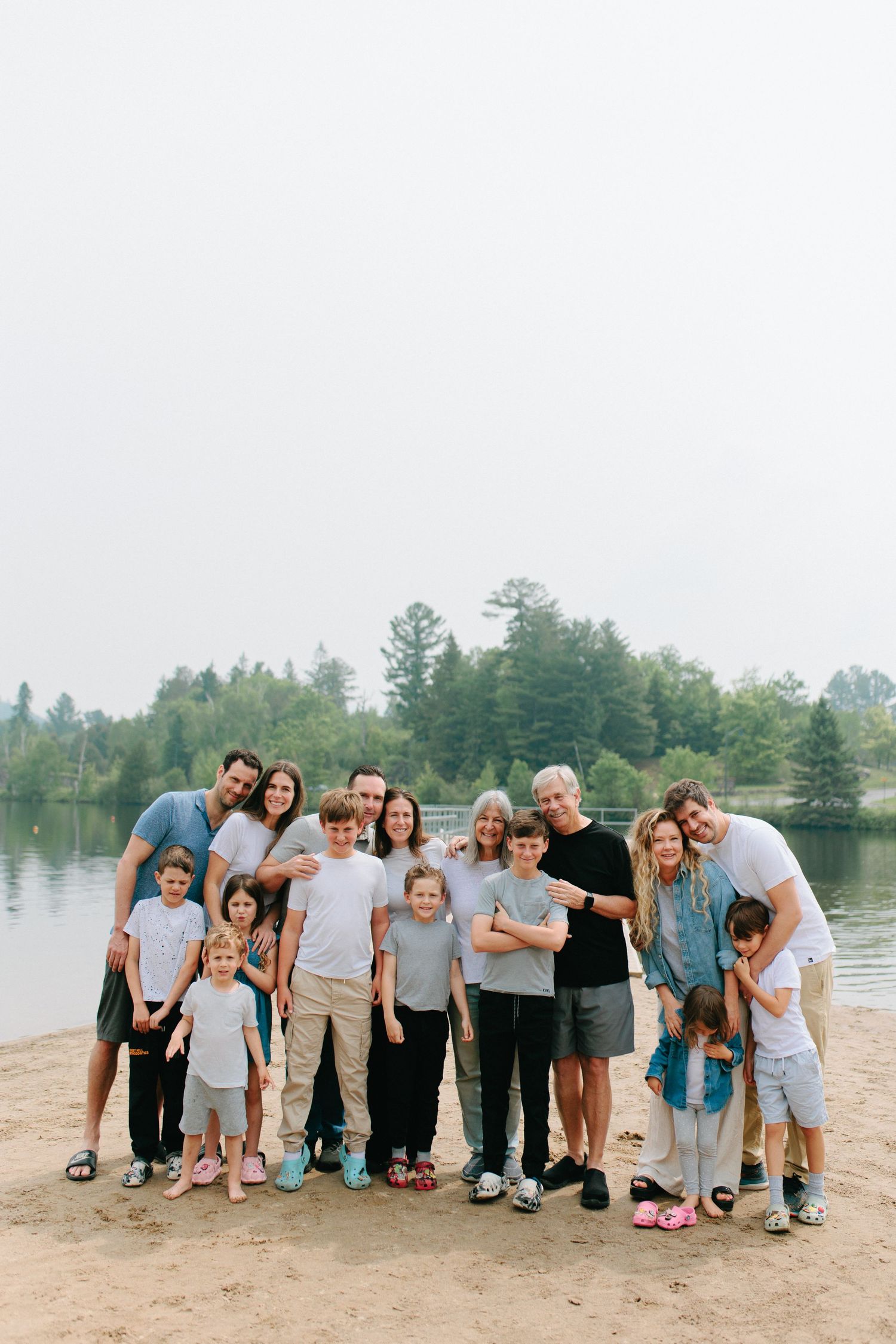A group of people standing together on a sandy beach by a lake surrounded by evergreen trees on a cloudy day.