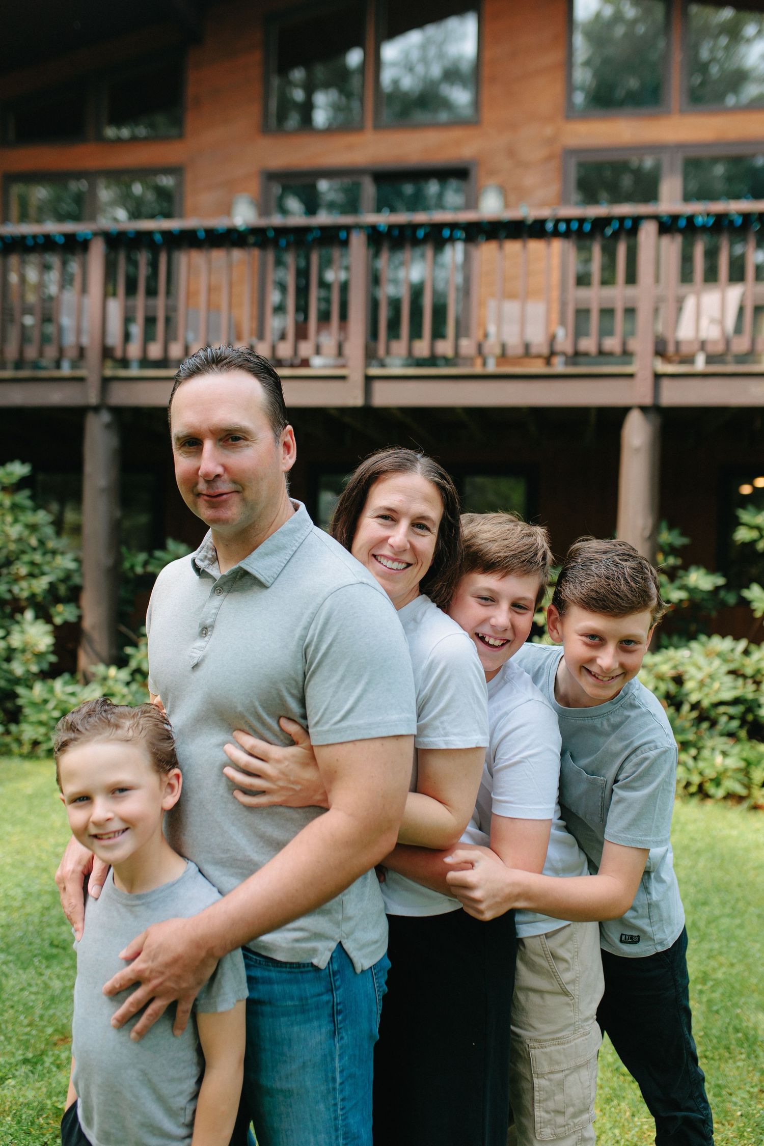 Family standing in line against a wooden deck house in the backyard, smiling and embracing each other.
