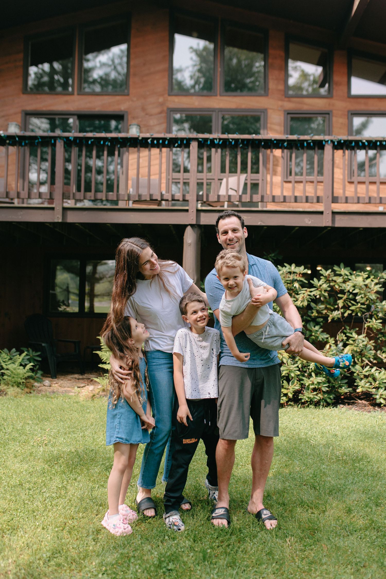 A family stands together smiling in front of a rustic wood cabin with a deck on a sunny summer day.