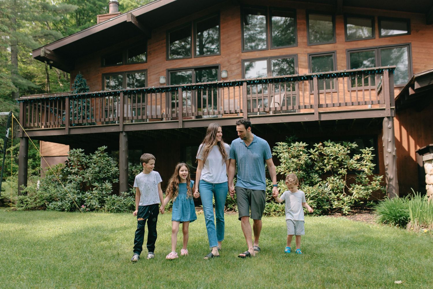 A scenic wooden cabin with a large deck surrounded by greenery as a family walks together across the front lawn.