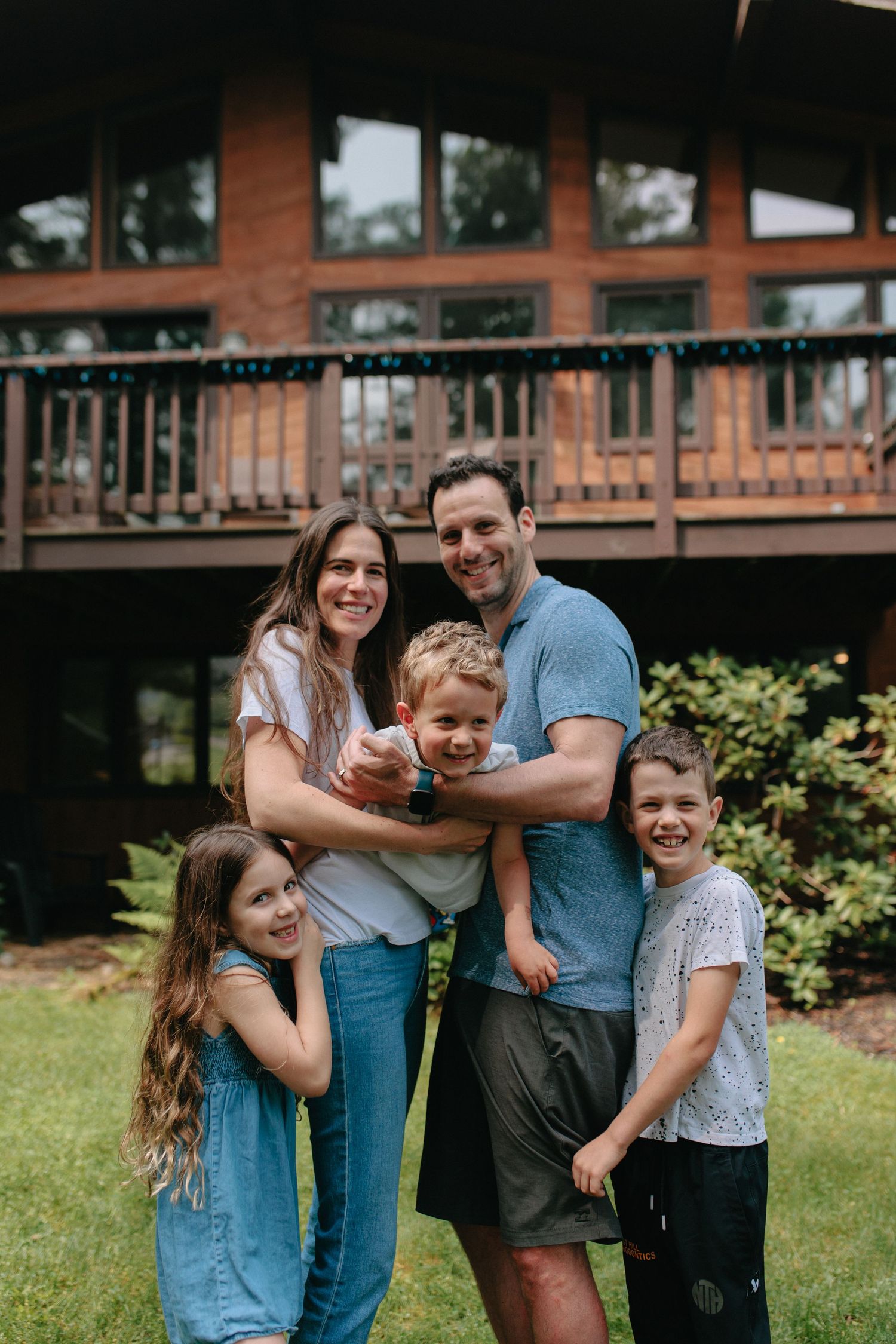 A cheerful family stands together on their lawn in front of a rustic cabin deck.