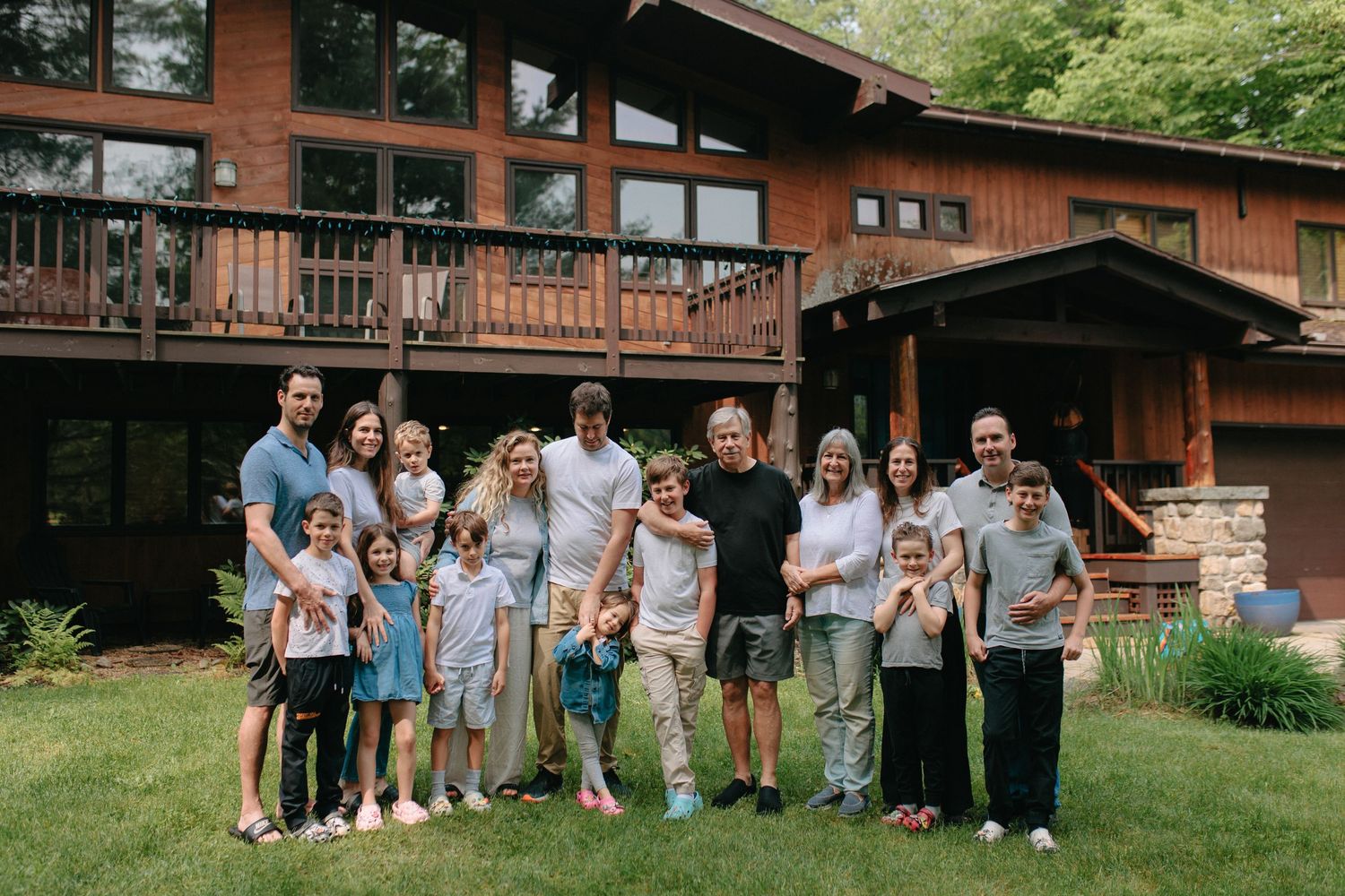 A group gathers outside a rustic wooden lodge with a deck and landscaped yard during summer.