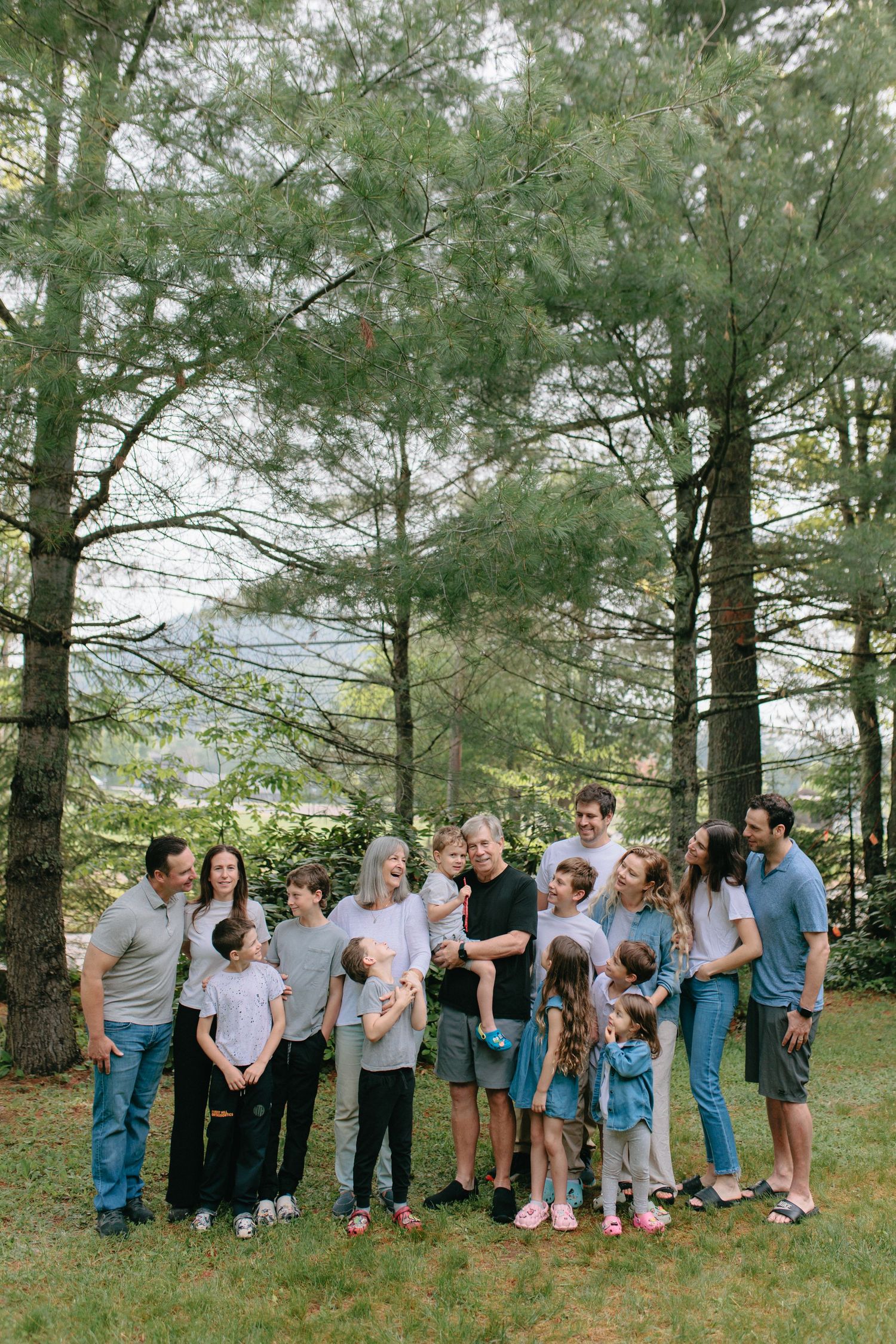 Large family group gathered outdoors in a pine forest setting wearing casual clothing for a portrait session.