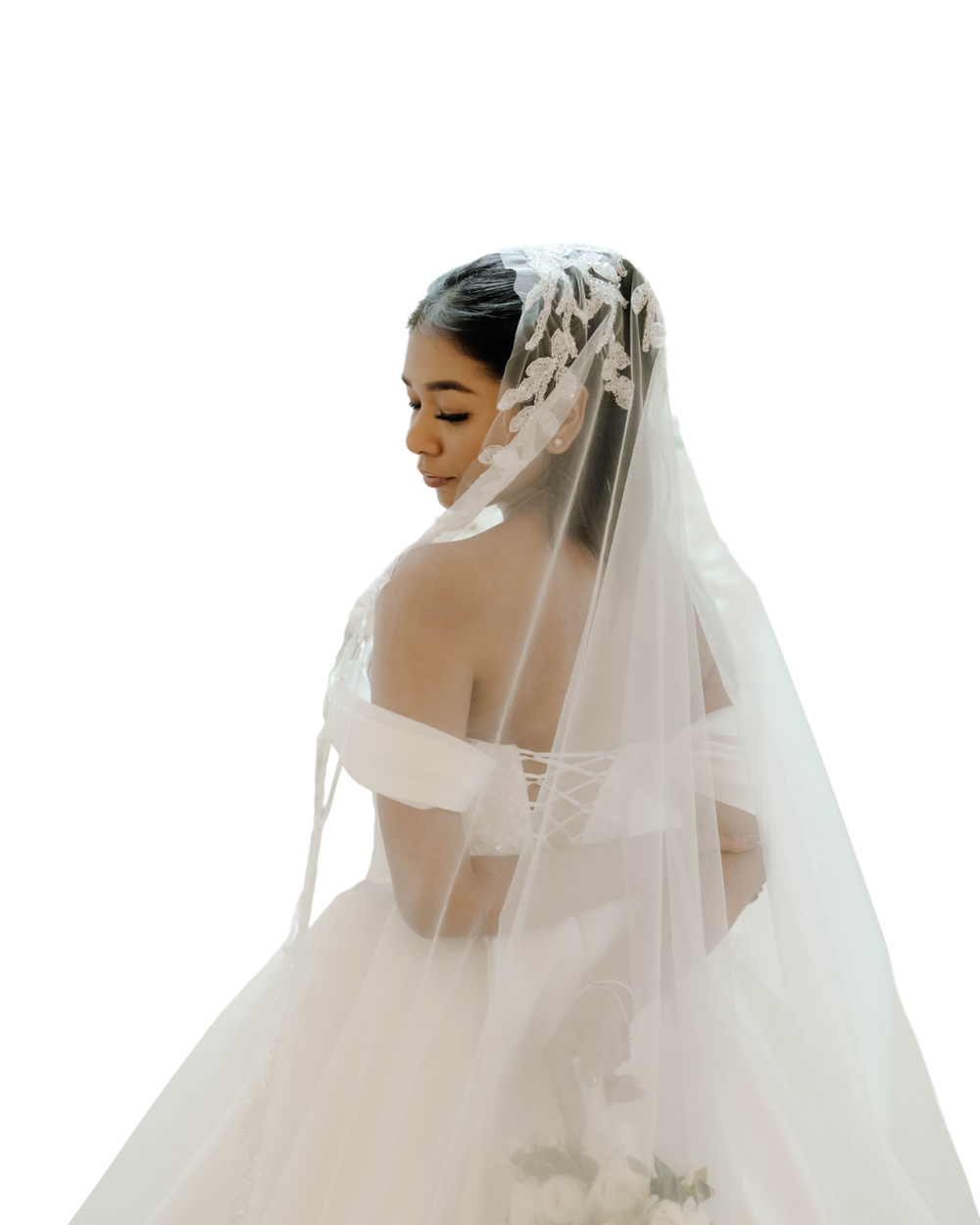 Side profile view of a bride wearing an elegant off-shoulder wedding gown and flowing sheer veil against white background.