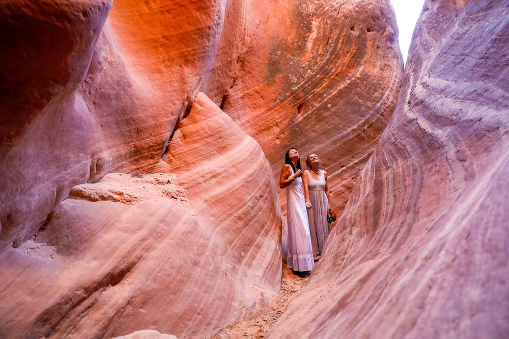 Two women in flowing dresses pose inside a stunning red rock slot canyon with swirling sandstone walls.