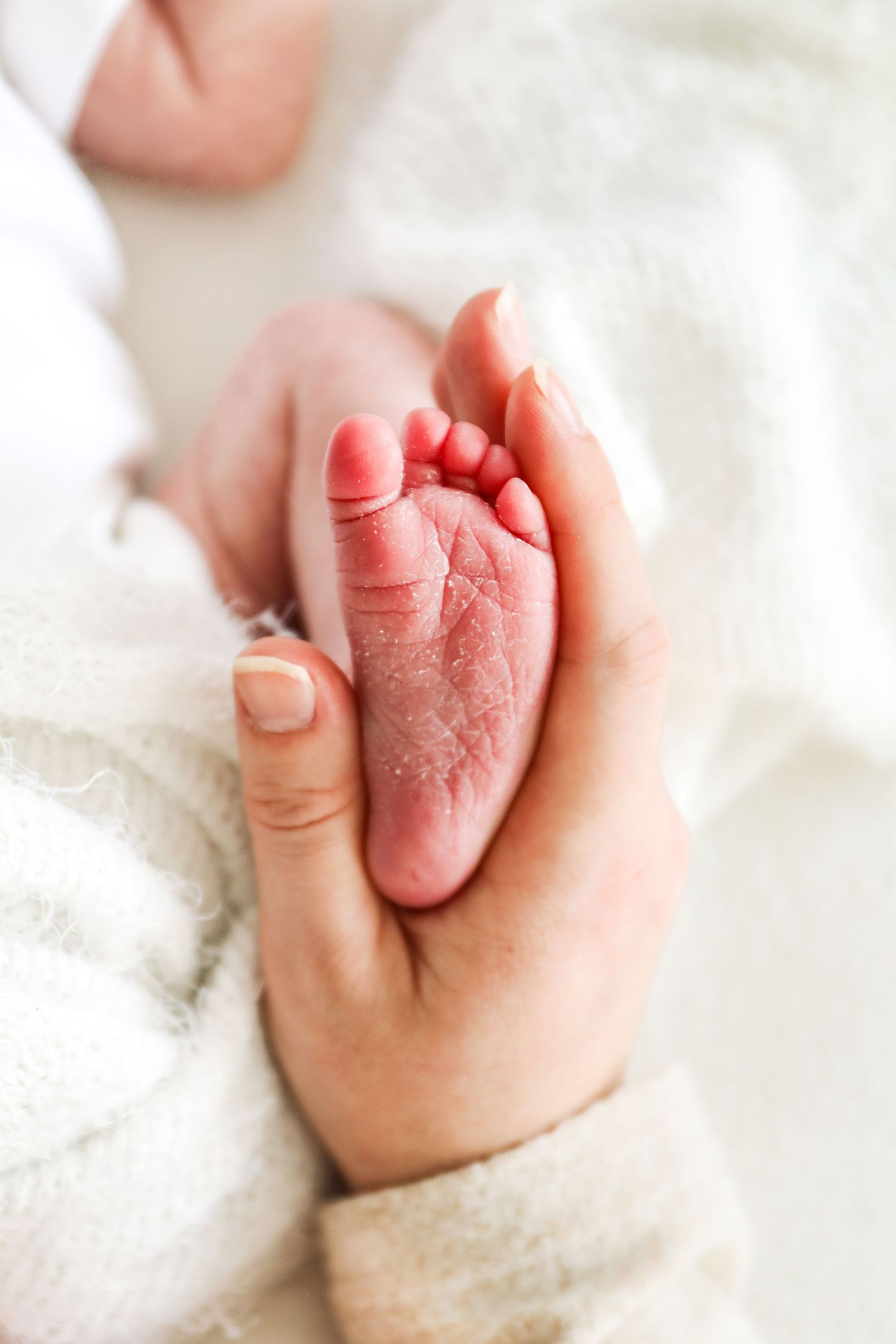 Adult hand gently holding tiny newborn baby foot against soft white blanket background, during Newborn shoot in Nottingham