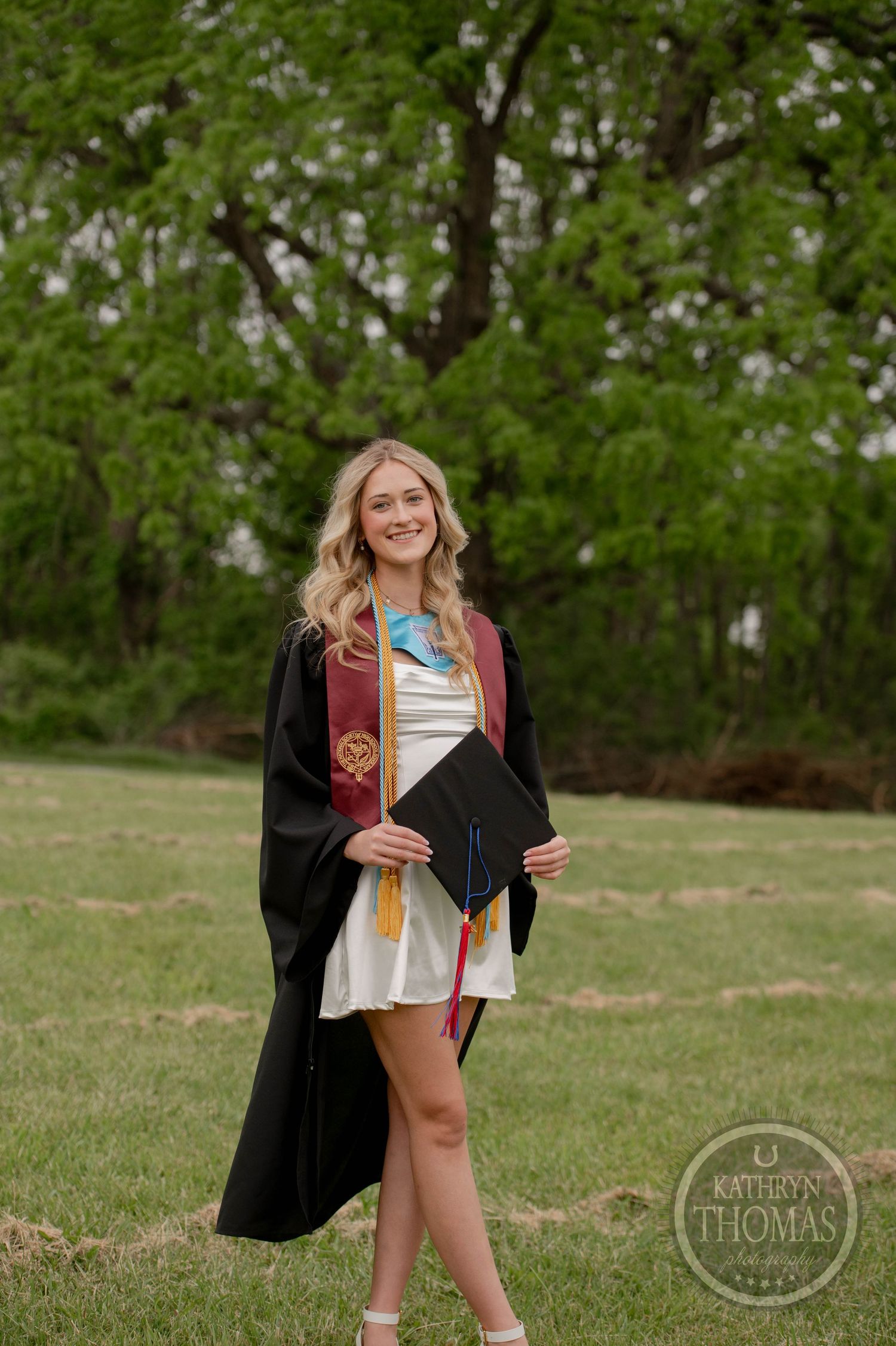 College graduate in black graduation gown poses with diploma outdoors against green trees on sunny spring day.