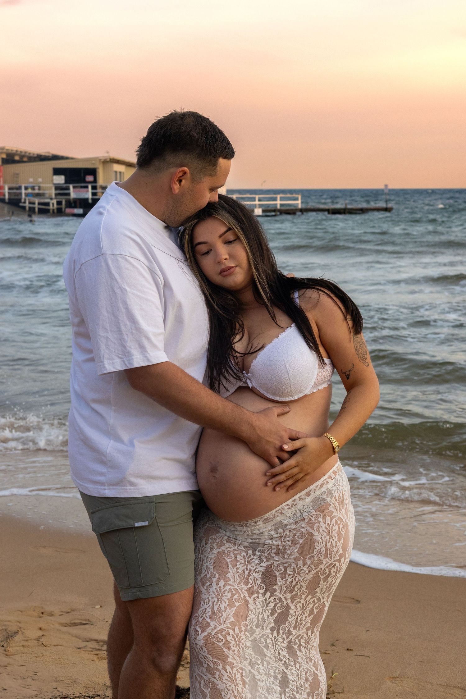 A couple embraces on the beach at sunset, the man holding the woman’s pregnant belly as waves roll gently behind them.