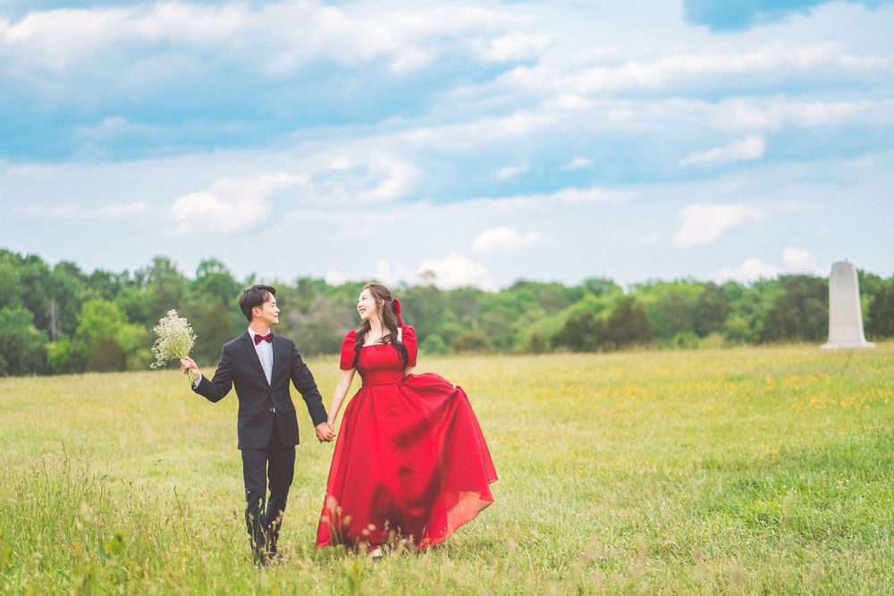 Couple walking through a sunny meadow, with the person in a flowing red dress and their partner in a black tuxedo.