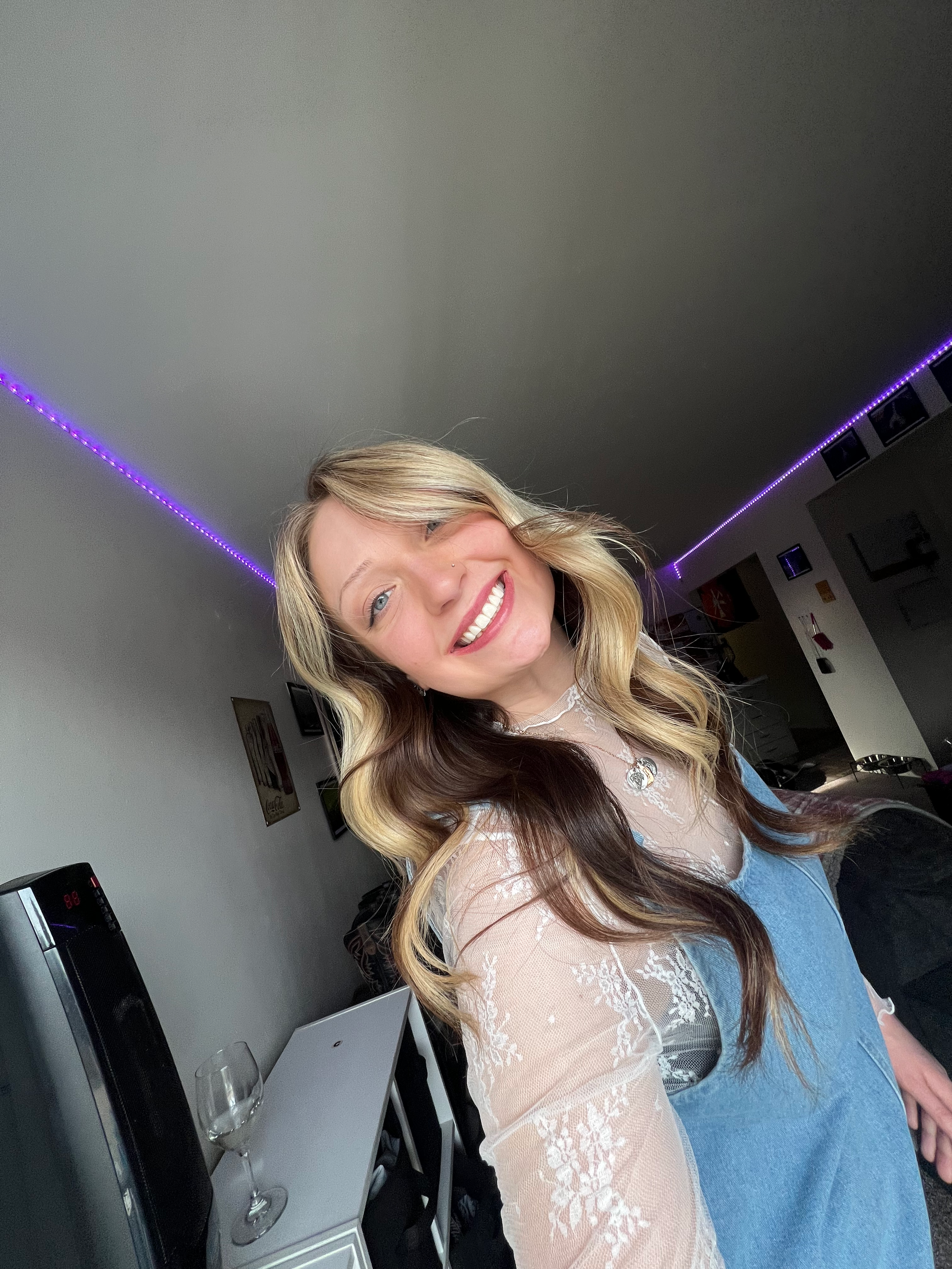 A person with long wavy blonde and brown hair wearing a light colored sweater smiles brightly in a selfie.