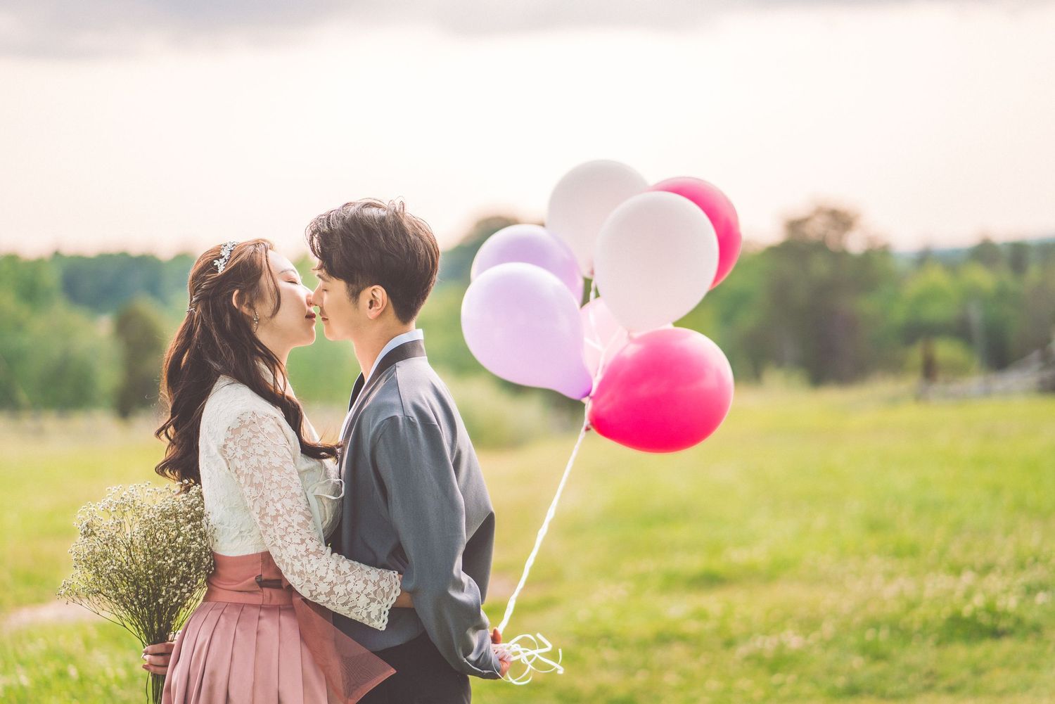 Romantic couple sharing a tender moment in a meadow at sunset with pink and white balloons.