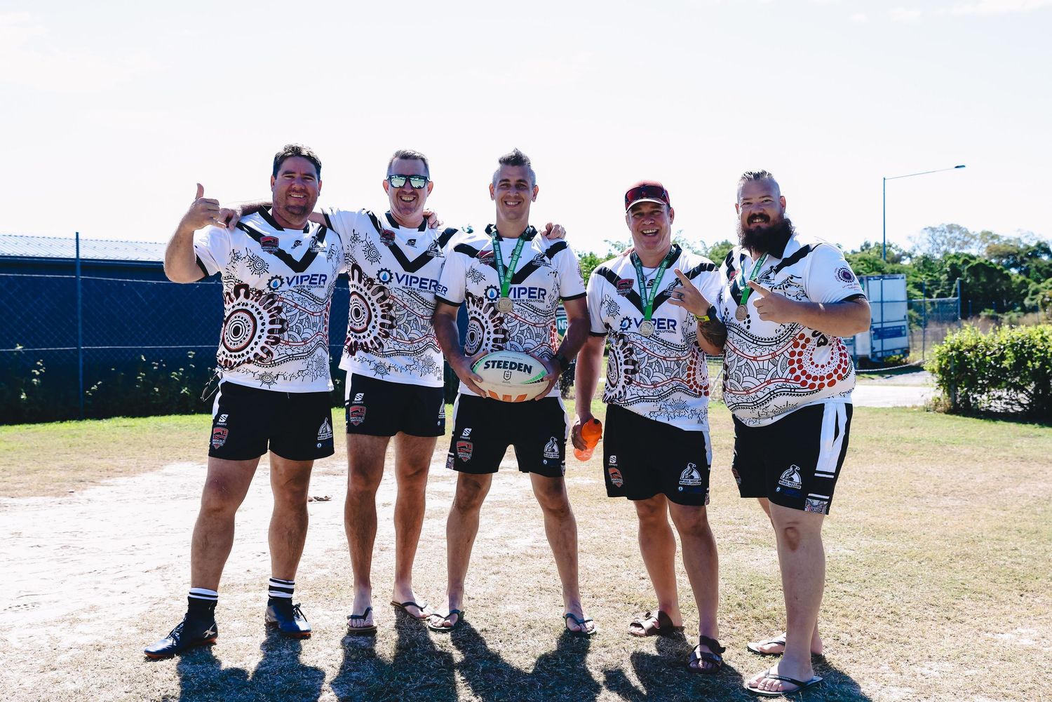 Rugby league players pose together wearing Indigenous design jerseys on a sunny field.