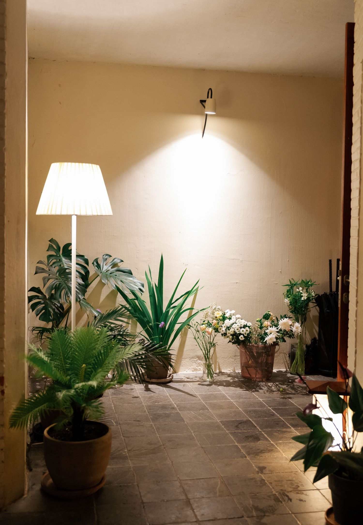 Indoor corner with potted tropical plants and decorative lighting featuring a floor lamp and wall-mounted sconce on gray tiles.