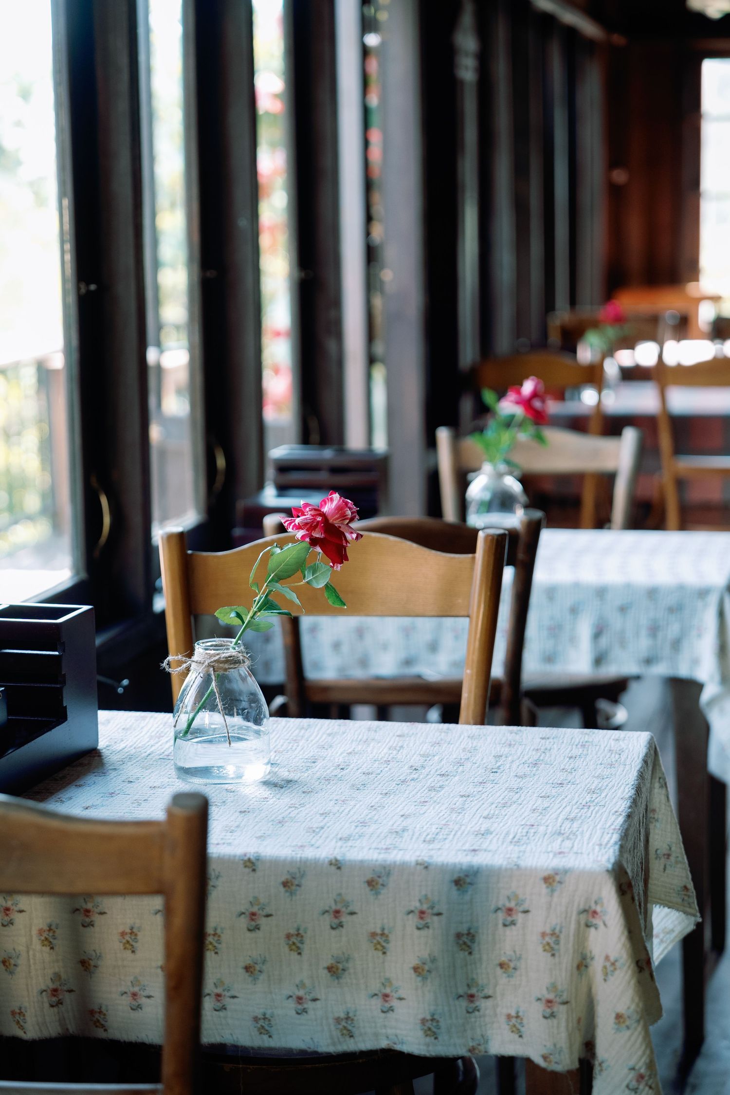 A rustic restaurant interior with wooden chairs and floral tablecloths, featuring a small glass vase with pink flowers.