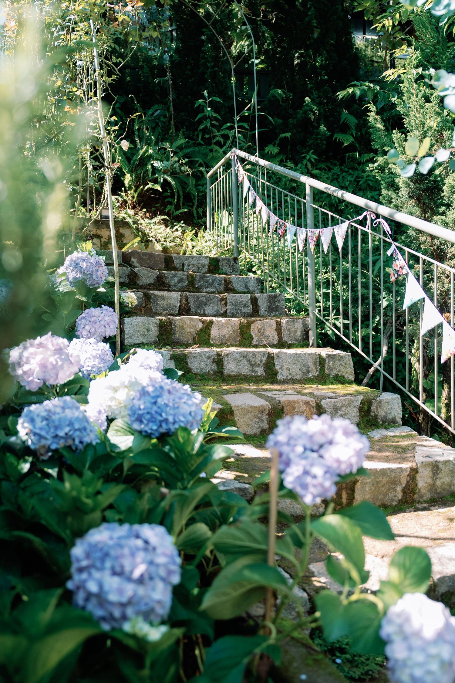 Stone garden steps with white metal railing surrounded by blooming blue hydrangea flowers in a lush garden setting.
