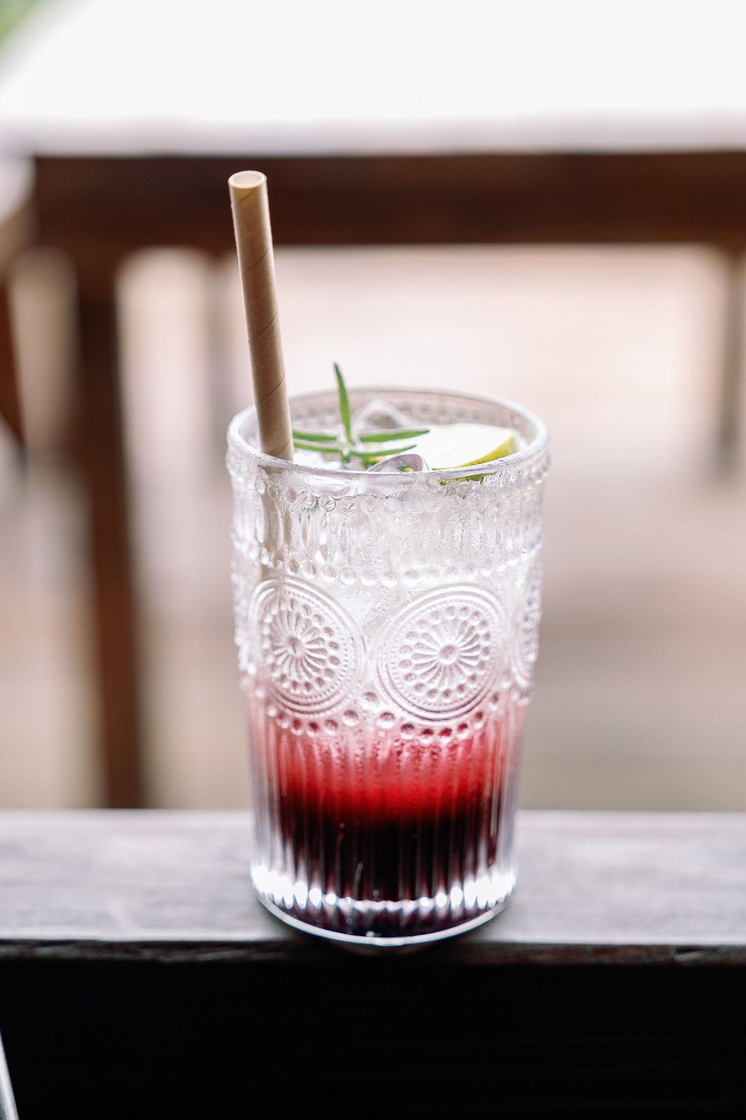 A layered red and dark cocktail served in an ornate vintage glass with a bamboo straw and herb garnish.