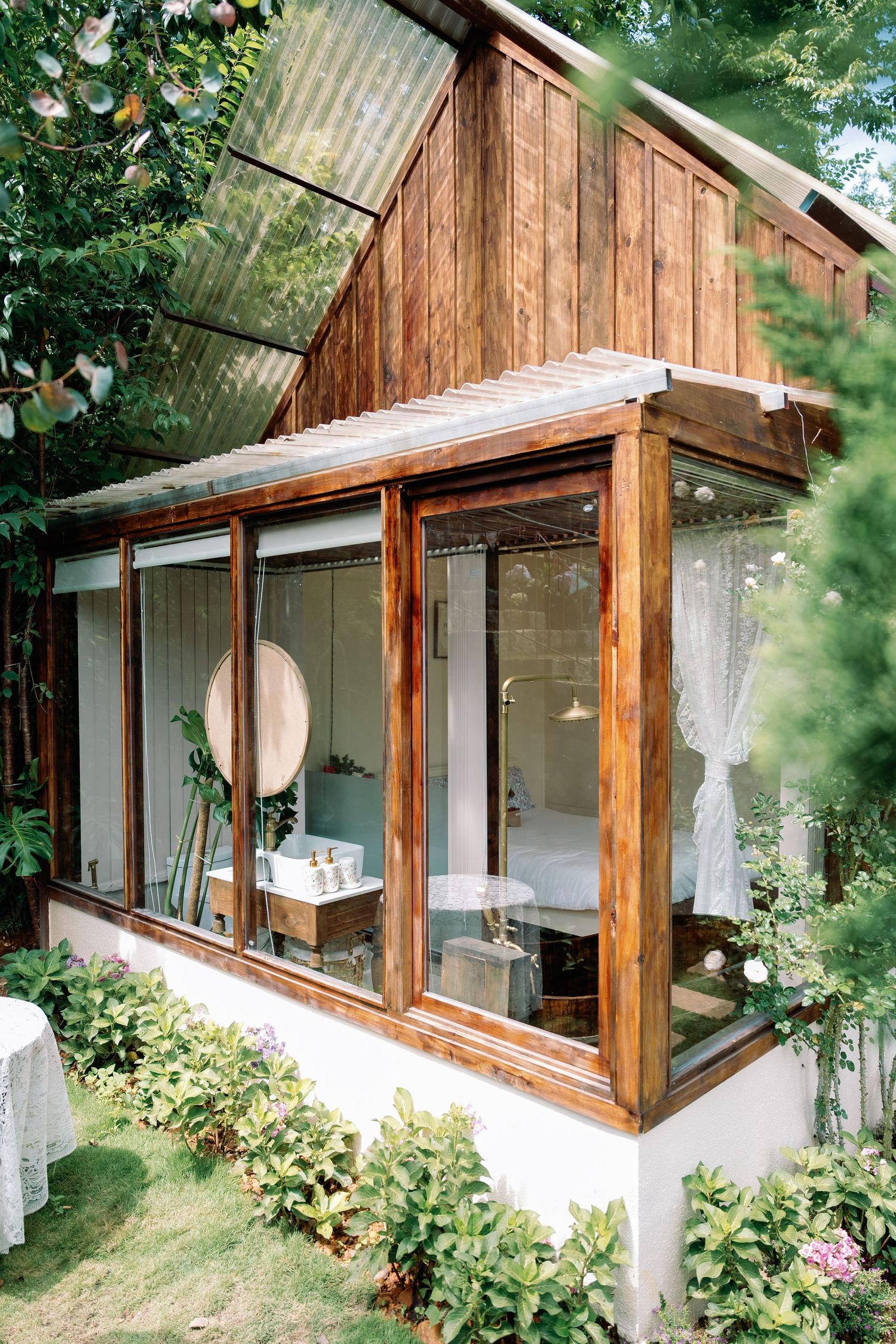 A rustic wooden cottage with large windows and white walls features climbing roses and greenery along its exterior.