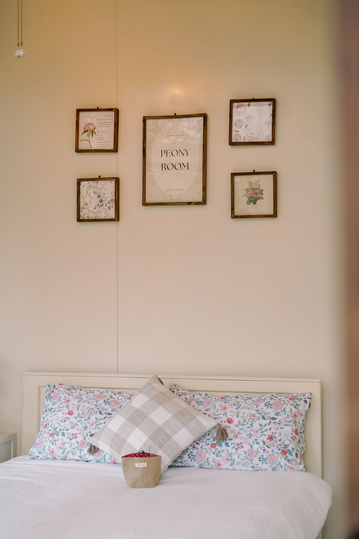 Cozy bedroom interior with floral pillowcases, gray checkered pillow, and framed botanical artwork on beige wall.