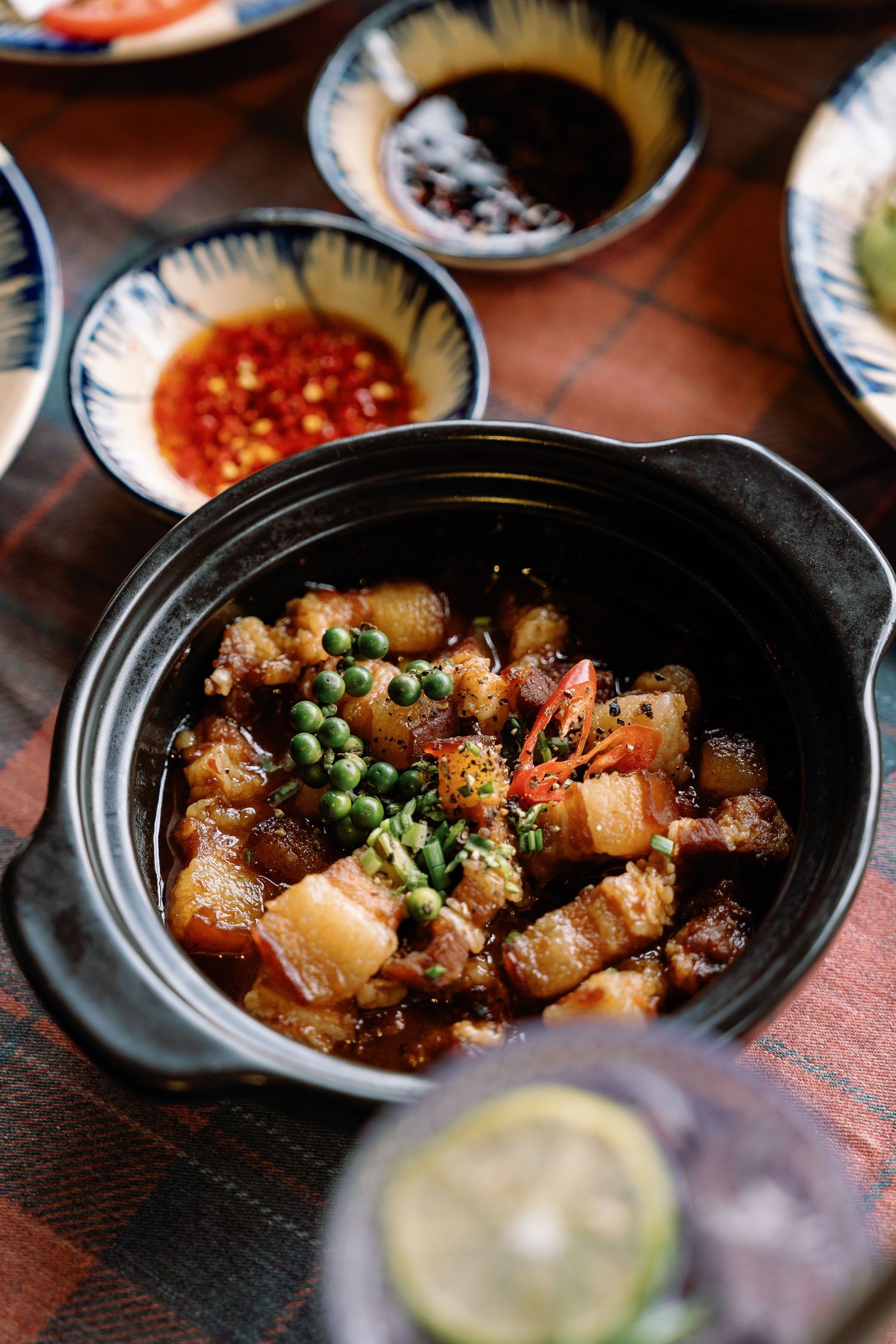 Clay pot dish filled with braised pork belly and green peas with dipping sauces in small bowls on a rustic red tablecloth.