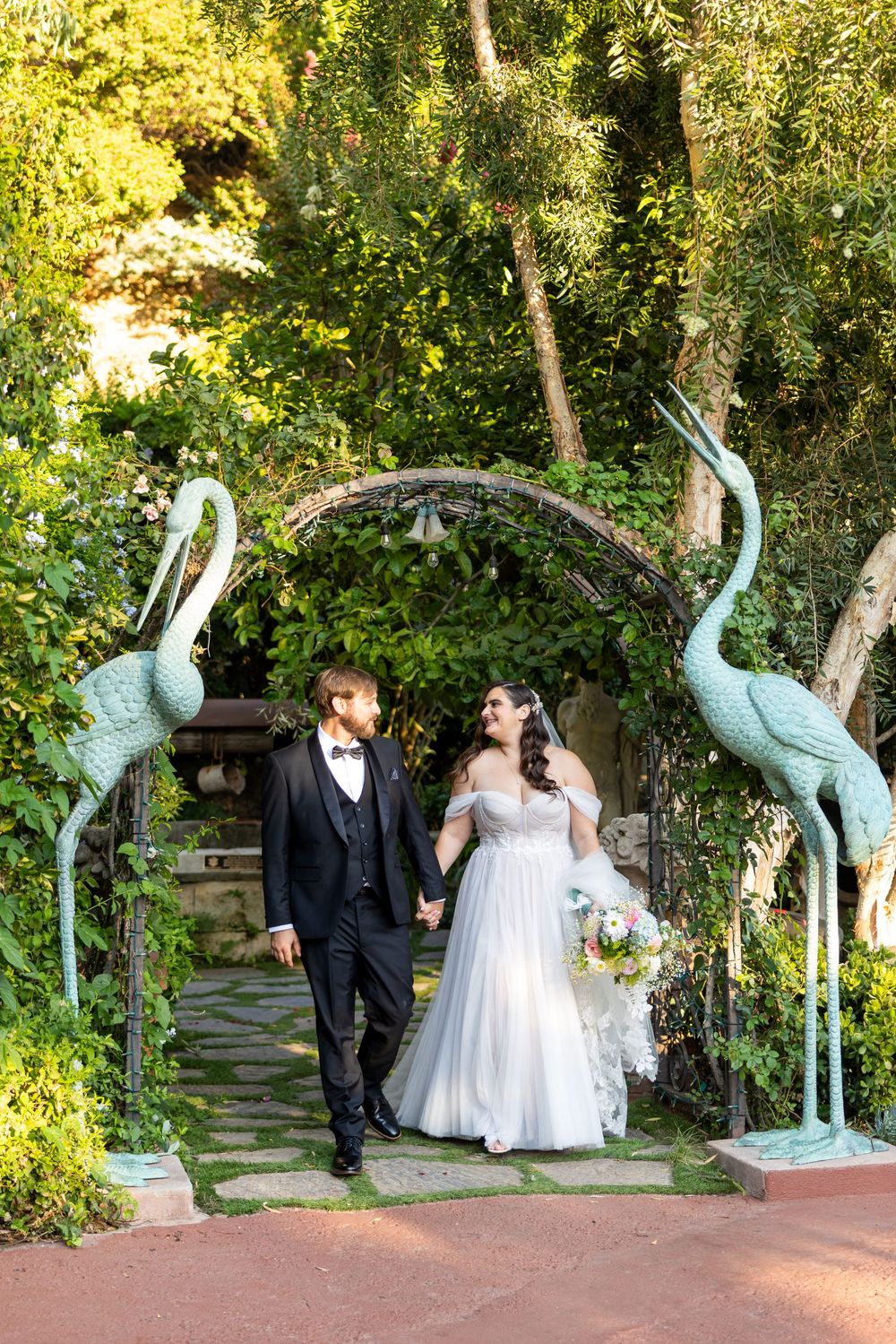 Wedding couple walk through garden archway flanked by decorative blue crane statues on a stone pathway.