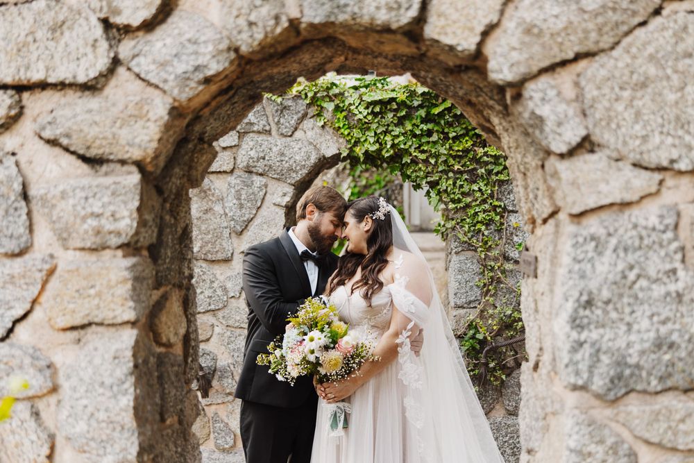 A romantic couple embraces in a stone archway adorned with climbing greenery while holding a wedding bouquet.