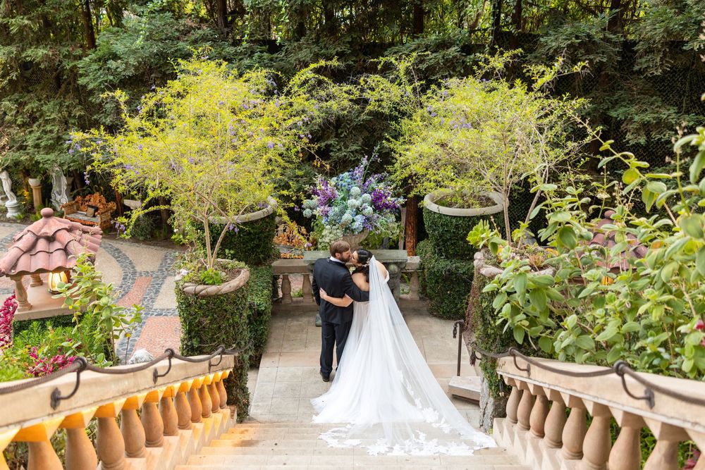 A couple walks down ornate stone stairs surrounded by lush gardens and potted plants at an outdoor venue.