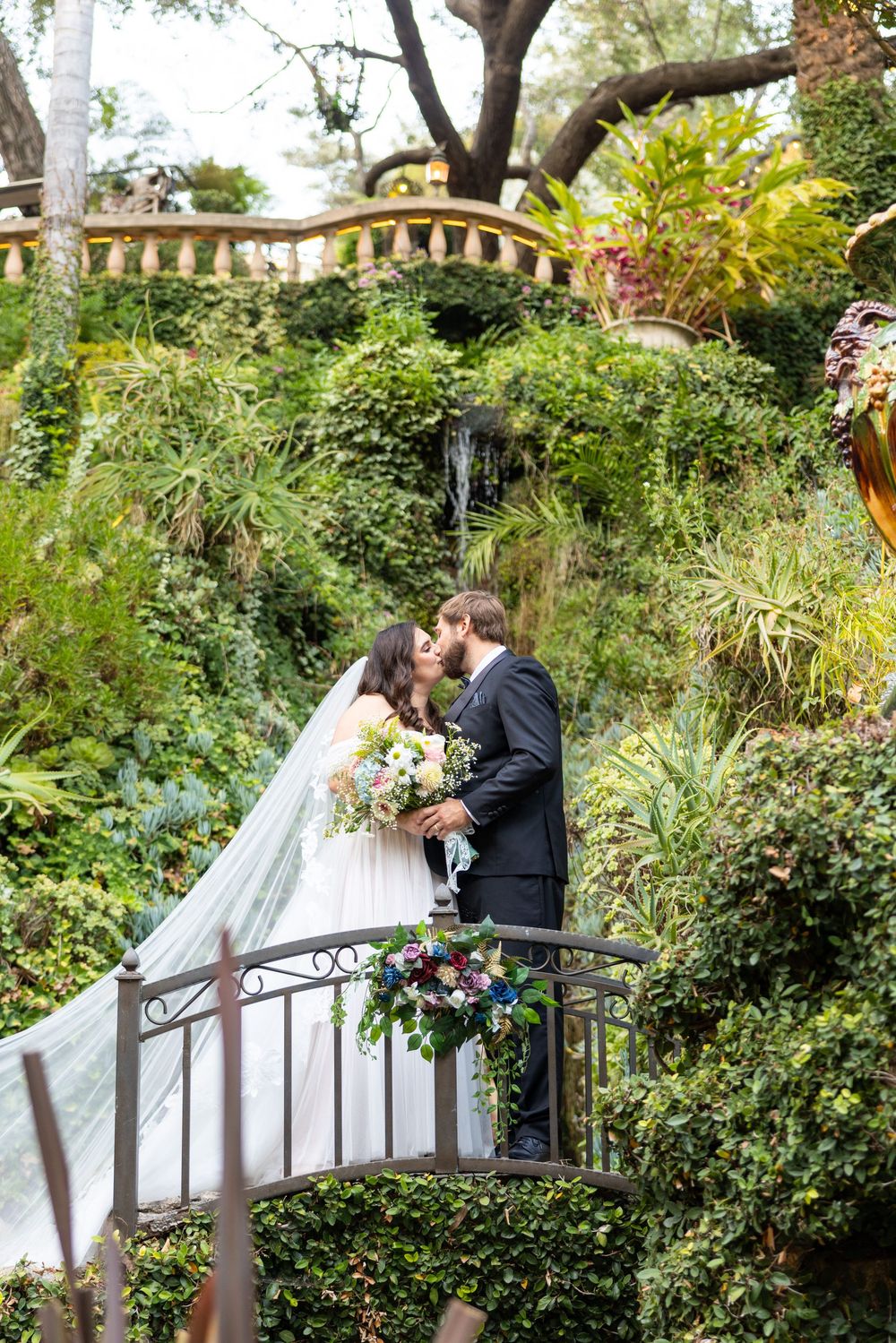 A couple shares a romantic moment on a garden bridge surrounded by lush greenery and flowers in an elegant outdoor setting.