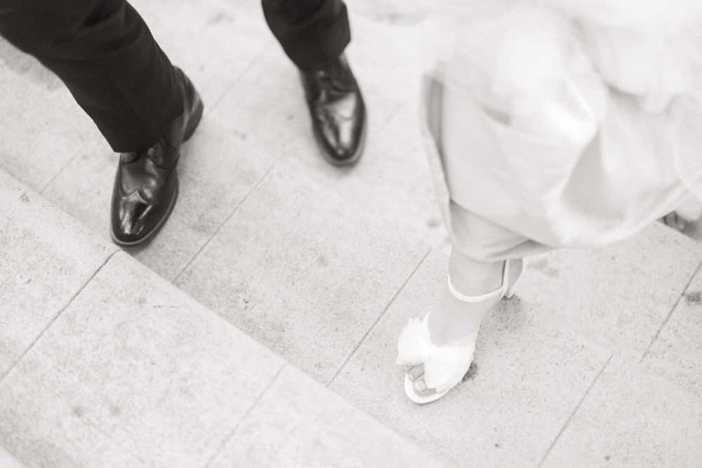 Black dress shoes and white bridal sandals stand side by side on a light colored floor.