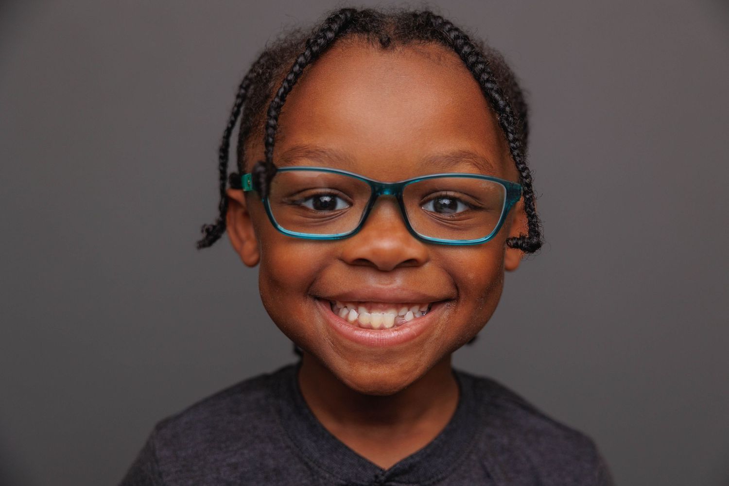 Portrait of a young student wearing glasses with braided hair and a bright smile against a gray background.