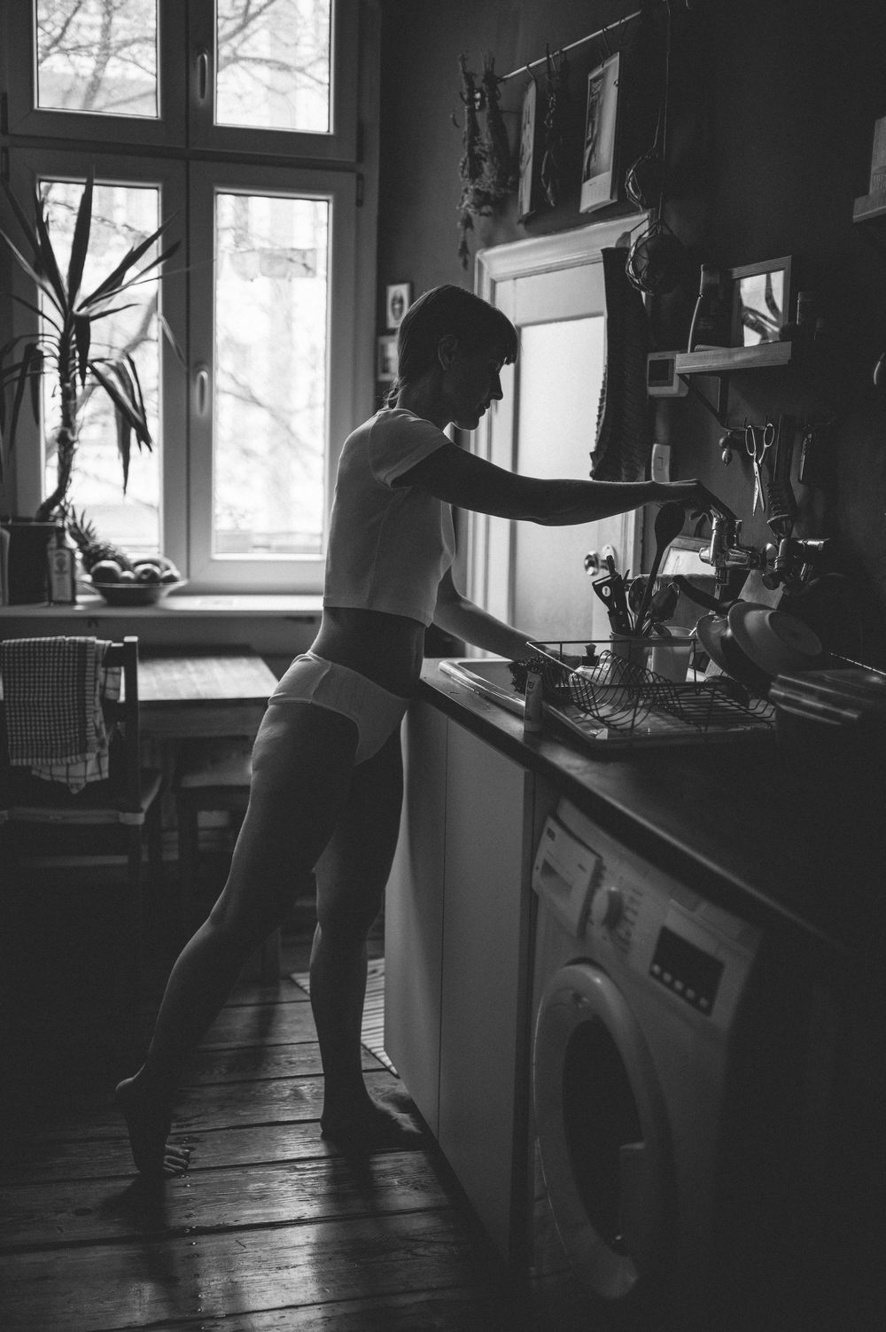 Black and white photo of a silhouetted figure working in a vintage kitchen interior with a window and washer.