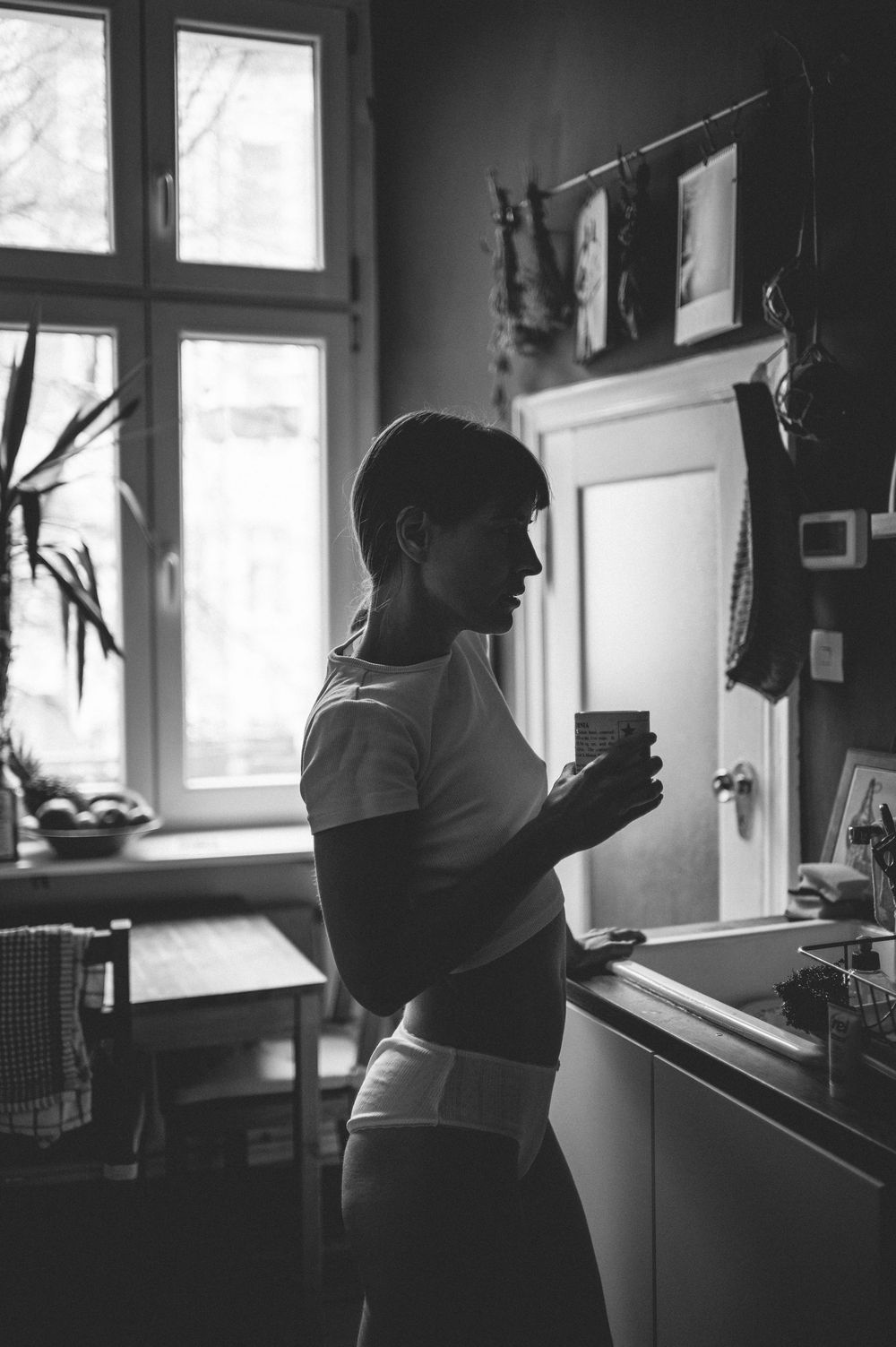 Black and white photograph of a silhouette holding a cup near a bright window in a kitchen setting.