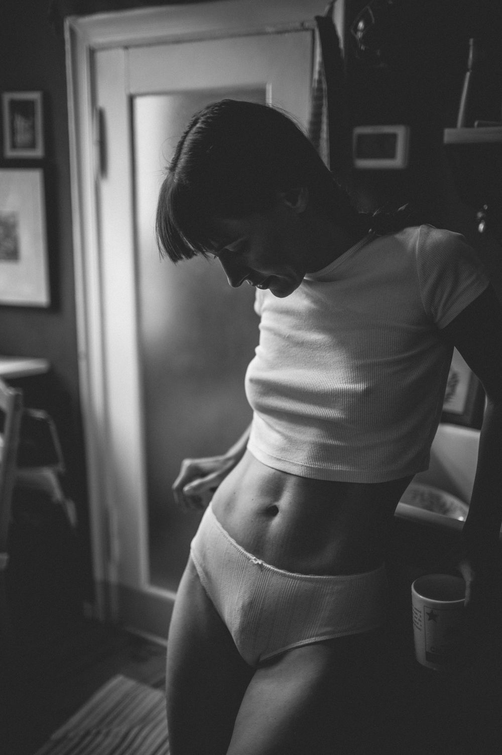 Artistic black and white photo showing a silhouette of someone in a cropped white tank top and underwear in a dimly lit room.