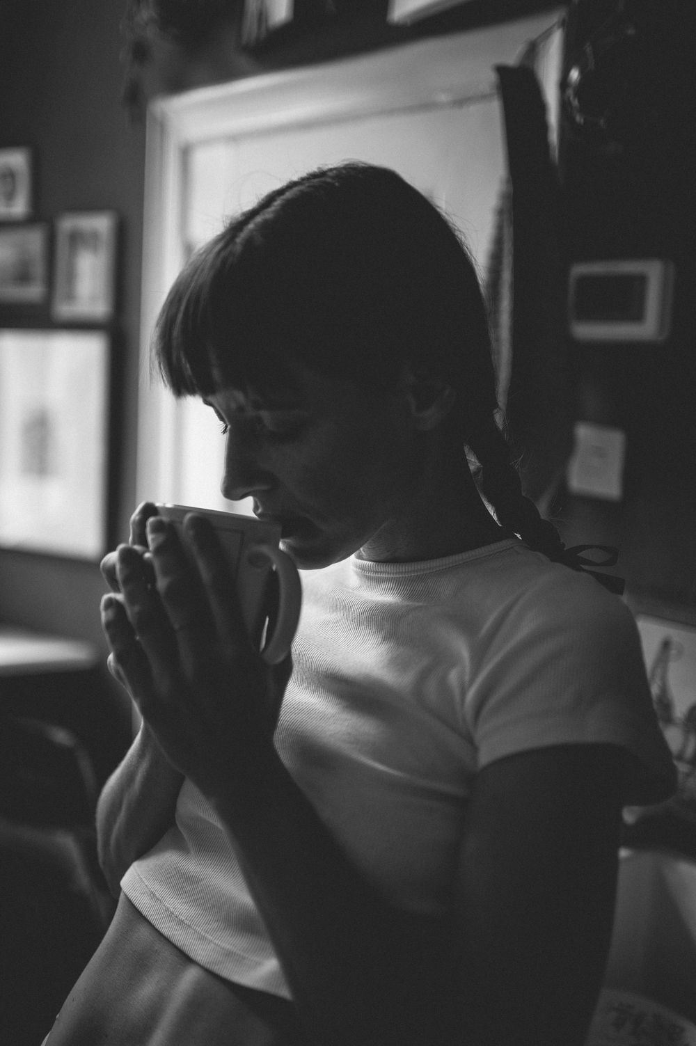 A black and white portrait showing a silhouette holding a steaming mug in a moody indoor setting with framed art on the wall.