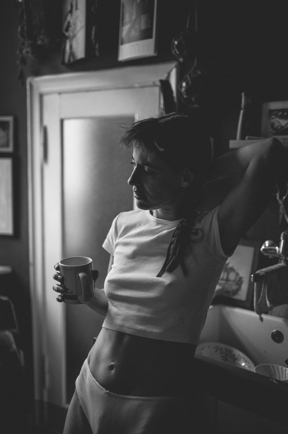 Black and white photograph of a person in a white t-shirt drinking from a mug in a dimly lit kitchen interior.