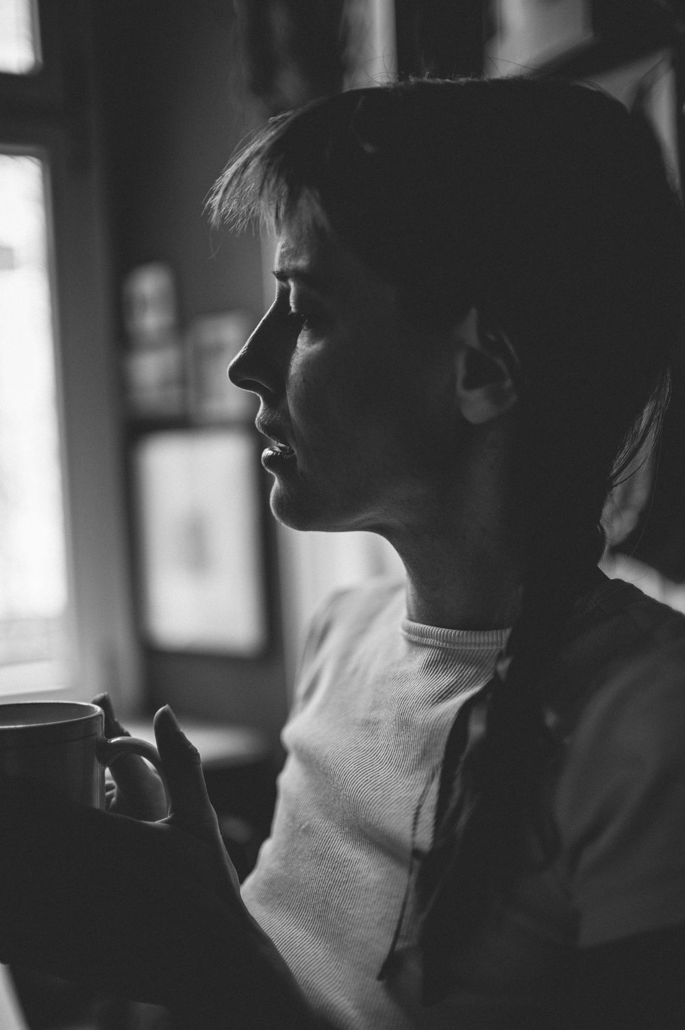 Black and white profile portrait in moody lighting shows a figure in a white t-shirt gazing out a window.