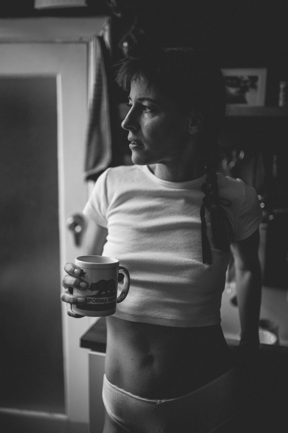 Black and white photo of someone holding a coffee mug while wearing a cropped white t-shirt in a dimly lit space.