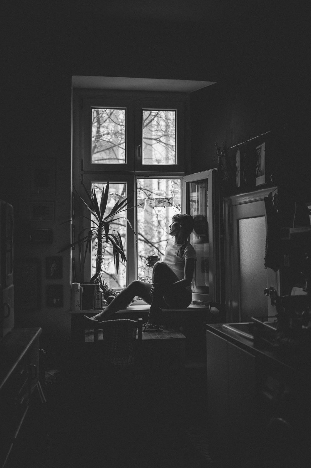 Atmospheric black and white photo of a silhouette sitting by a tall window with a houseplant in a dark interior.